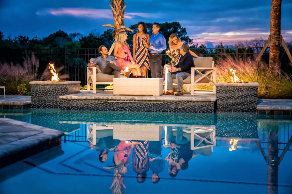 Group Of Three Couples Sitting At Night By a Pool Next To A Pair Of Fire Pits.